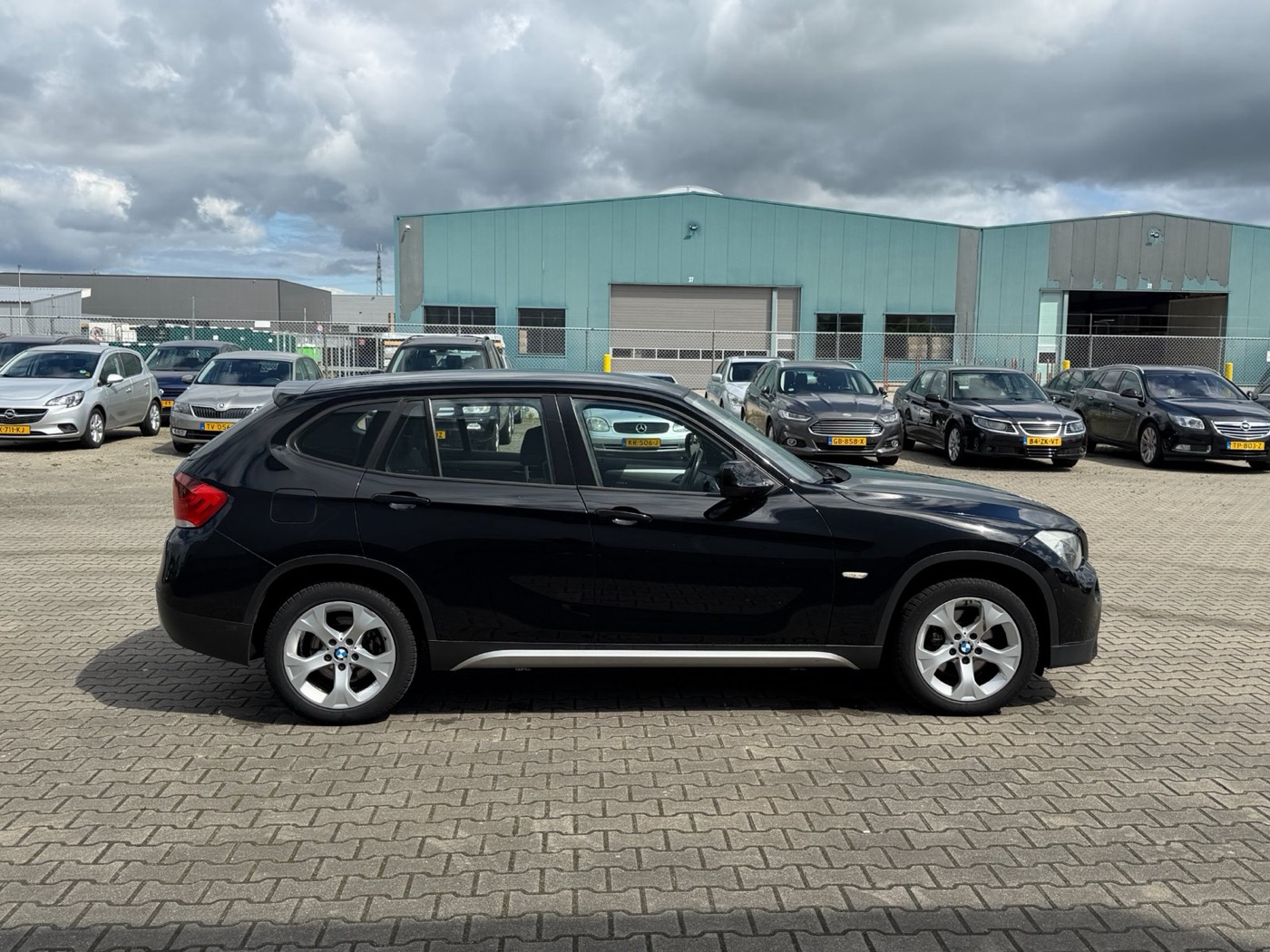 Zwarte BMW X1 van opzij op een dealerterrein met bewolkte lucht en een rij geparkeerde auto's erachter